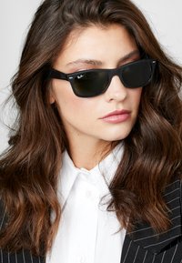 Occhiali da sole in plastica nera con lenti scure. Presentano dettagli in argento e rivetti in metallo rotondi. Indossati con una camicia bianca e un blazer a righe.