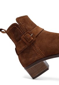 Stivaletto in suede marrone con punta arrotondata, una cinghia decorativa con fibbia e un tacco impilato. Presenta dettagli di cucitura lungo le cuciture.