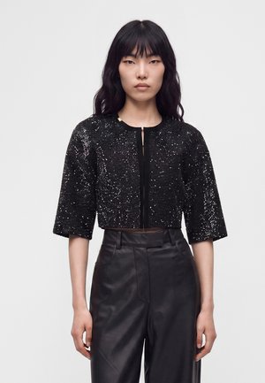 Frau mit langen schwarzen Haaren, die eine schwarze kurze Paillettenjacke und hochtaillierte schwarze Lederhose trägt, steht vor einem schlichten weißen Hintergrund.