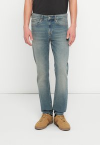 Vaqueros de mezclilla azul claro con corte de pierna recta y un sutil lavado, combinados con zapatillas de ante color tostado. La cintura tiene trabillas para cinturón y herrajes metálicos.