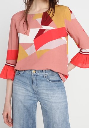 Vrouw die een kleurrijke blouse met geometrisch patroon en 3/4 gerafelde mouwen draagt, gecombineerd met lichtblauwe high-waist jeans.