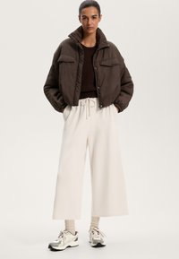 Barna, rövid pufikabát, két elülső zsebbel, laza illeszkedésű krémszínű nadrággal és több színű sportcipővel párosítva. Egyszínű háttér.