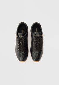 Ζευγάρι μαύρα δερμάτινα sneakers Tommy Hilfiger με μαύρες κορδόνες και καφέ λάστιχο, απεικονιζόμενο από πάνω σε λευκό φόντο.