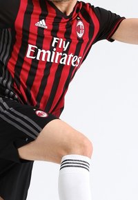 Punainen ja musta raidallinen jalkapalloilupaita, jossa on pyöreä kaula-aukko, Adidas-logo ja seuran emblem. Mustat shortsit, joissa on harmaat sivuraidat. Valkoiset sukat, joissa on mustat raidat.