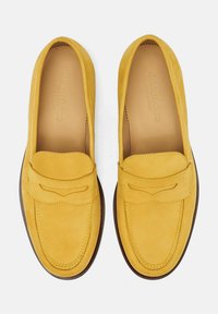 Mocasines de ante amarillo con una costura decorativa en el frente y una correa decorativa, que presentan una textura suave y una suela de goma marrón.