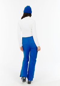 Blauwe skibroek met een aansluitende taille en uitlopende pijpen, gecombineerd met een witte lange mouwtop en een blauwe gebreide beanie. Gestucte stof met ritsen.