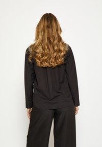 Čierna košeľa s dlhými rukávmi a hladkým materiálom, s gombíkovými manžetami a zaobleným lemom. Vlnité vlasy padajú dolu po chrbte.