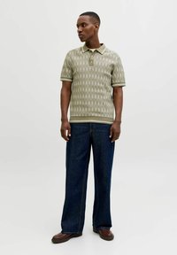 Mann trägt grünes, gemustertes, kurzärmeliges Polo, dunkle Jeans mit weitem Bein und braune Schuhe, steht vor hellem, einfarbigem Hintergrund.