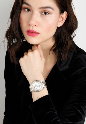 Nuori nainen tummilla hiuksilla, musta toppi päällään, nojaa leukaansa käteensä ja esittelee hopeanväristä rannekelloa, jossa on valkoinen kellotaulu.