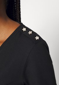 Zwarte V-hals top met drie zilveren ster embellishments op de schouder, gemaakt van zachte stof met een gladde textuur.