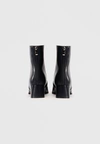 Botas de cuero negras hasta el tobillo con una textura suave, tacones cuadrados y cremalleras en la parte trasera. El diseño presenta una silueta limpia y minimalista.