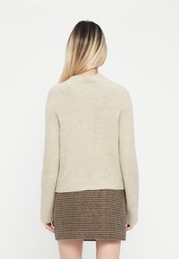 Beige neulemekko, jossa on ribbireuna ja pitkät hihat, käytössä ruskean houndstooth-kuvioidun hameen päällä. Kangas näyttää pehmeältä ja tekstuuriltaan rikkalelta.