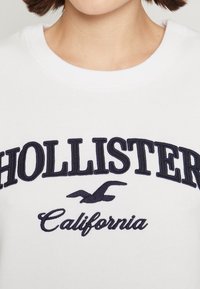 Vit sweatshirt med marinblå broderad text "HOLLISTER" och Kalifornien-design. Har rund halsringning och mjukt tyg.