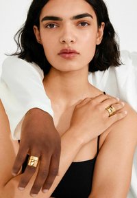 Kuldrenkad kujundusega sõrmused kaetud käte peal, mis embavad paljast õlga, taustal hele valgus ja pehme kangas.