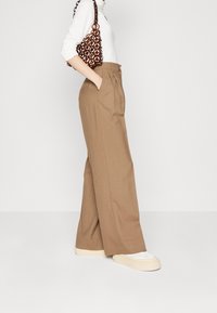 Wide-leg brown trousers made from textured fabric, featuring side pockets and high waist. Paired with a white top and chunky sneakers.