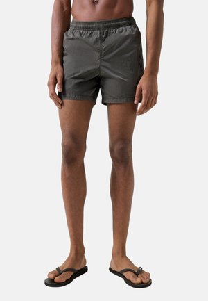 Mann trägt dunkelgraue Shorts mit elastischem Bund und schwarze Flip-Flops, steht vor schlichtem hellem Hintergrund.