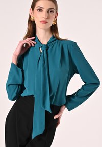 Blusa di seta turchese con colletto con laccio, maniche lunghe e dettagli plissettati; presenta dettagli con bottoni sui polsini e una vestibilità rilassata.