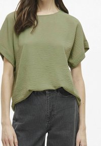 Top verde oliva con texture, maniche corte e una vestibilità ampia, abbinato a jeans grigio scuro. Il tessuto sembra leggero e leggermente stropicciato.