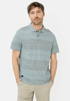 Mężczyzna o krótkich, brązowych włosach, ubrany w jasnoniebieską, pasiastą koszulkę polo i beżowe spodnie, stoi z rękami w kieszeniach na jednolitym tle.