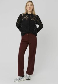 Blouse en dentelle noire avec des manches bouffantes, associée à un pantalon large bordeaux. Le mannequin porte des baskets argentées, mettant en valeur le tissu texturé et la coupe décontractée.