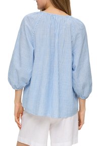Hellbluse mit gestreiftem Muster und luftigen Ärmeln, weicher Textur, rundem Ausschnitt und gerafften Details am Rücken. Kombiniert mit weißen Shorts.