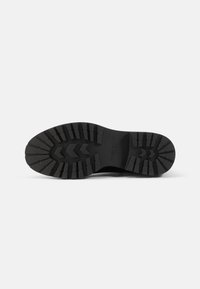 Semelle de chaussure noire avec des crampons profonds, des motifs en chevrons et rectangulaires pour la traction, et nom de la marque embossé près de la zone de la voûte plantaire.
