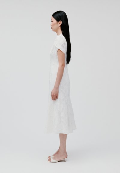 Abito bianco, strutturato, con maniche corte, lunghezza al ginocchio e motivi floreali. La modella indossa sandali con tacco aperto. Sfondo neutro.