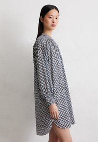Blau-weiß gemustertes Kleid mit langen Ärmeln, einem runden Ausschnitt und einem längeren Rückensaum. Enthält geometrische Formen in Gelb und Schwarz.
