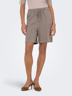 Vrouw die taupekleurige short met trekkoord draagt, beige top met kanten mouwen en bruine slingback pumps met hak, staand voor een effen achtergrond.