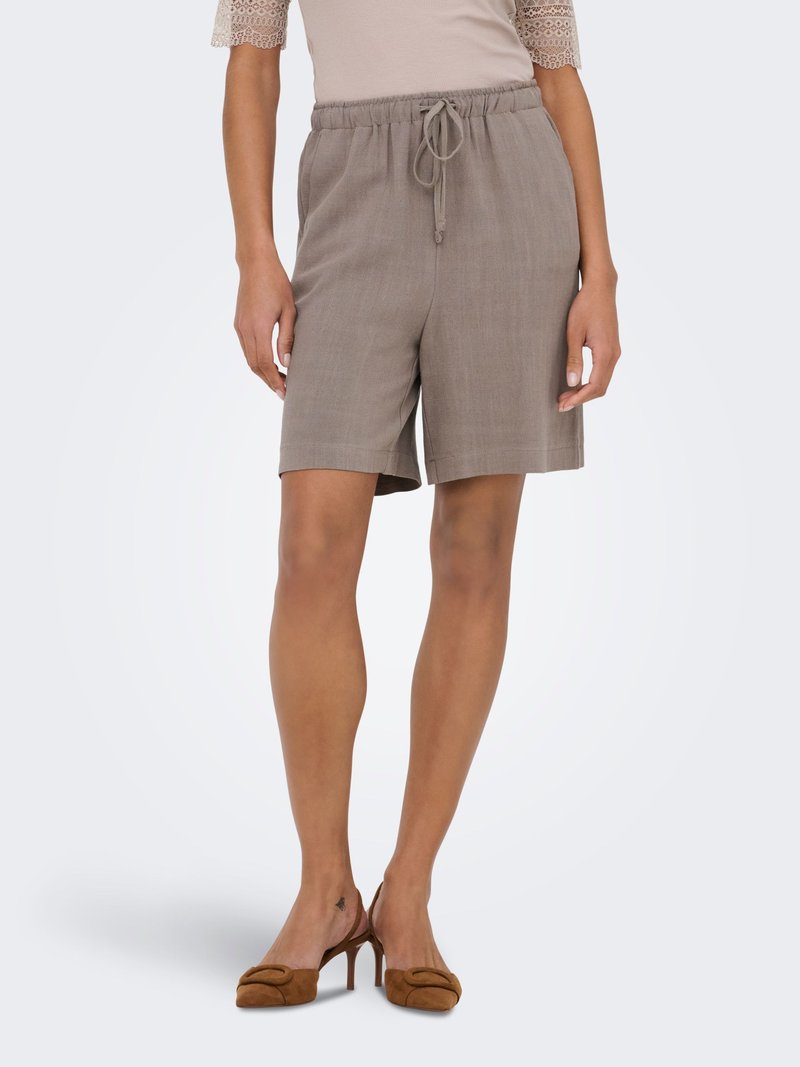 Femme portant un short taupe à cordon, un haut beige à manches en dentelle et des chaussures à talons slingback marron, debout devant un fond uni.