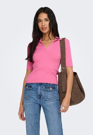 Kvinde med langt mørkt hår iført en lyserød ribbet polo-shirt og blå jeans, der bærer en stor brun ruskindtaske over skulderen.
