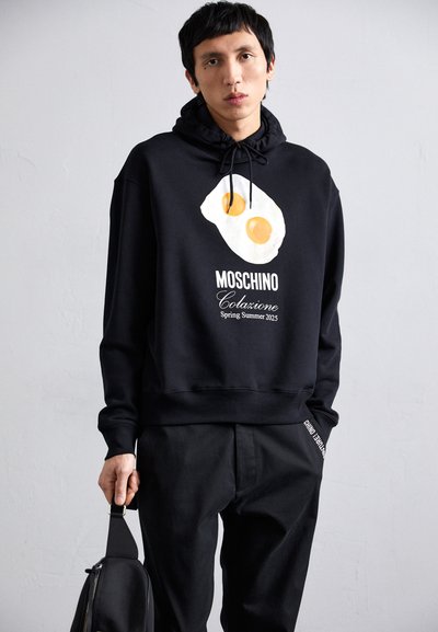 Schwarzer Hoodie mit einer Grafik von zwei Eiern, dem Text "Moschino Colazione" und einer Kordelzugkapuze. Getragen mit schwarzen Hosen; das Model hält eine schwarze Tasche.