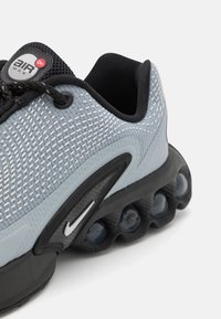 Zapatilla deportiva en gris y negro con parte superior de malla texturizada, prominente amortiguación Air Max y una suela suave y redondeada con bolsillos de aire visibles.