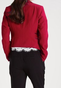 Femme portant un blazer rouge sur un haut en dentelle blanche et un pantalon noir, vue de dos avec de longs cheveux bruns.