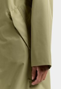 Wodoodporny płaszcz w oliwkowej zieleni o gładkiej fakturze; posiada widoczną kieszeń i subtelne logo na rękawie. Dopasowany krój ze prostymi rękawami.