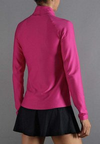 Vrouw in een gestructureerde roze top met lange mouwen en een korte zwarte plissékrok, gezien vanaf de achterkant tegen een grijze achtergrond.