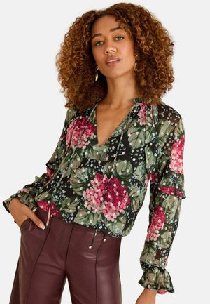 Bluse mit floralem Druck aus schwarzem, transparentem Stoff, verziert mit grünen Blättern und pinken/roten Blumen, mit Rüschensäumen und V-Ausschnitt. Kombiniert mit braunen Hosen.