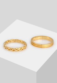 Due anelli d'oro su uno sfondo grigio chiaro. L'anello a sinistra presenta un design attorcigliato, mentre l'anello a destra ha bande lisce e sovrapposte.