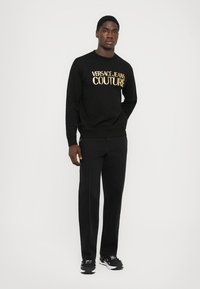 Črna majica s kapuco z napisom "Versace Jeans Couture" v zlatem tisku, v kombinaciji s črnimi hlačami z bočnimi šivi, oblečeno z črnimi športnimi čevlji.