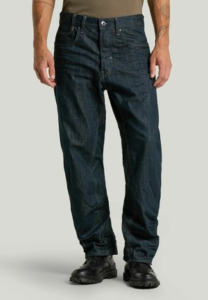 Man draagt donkerblauwe spijkerbroek en zwarte veterschoenen, staat met ontspannen armen langs zijn lichaam tegen een effen achtergrond.