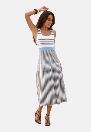 Femme souriante posant avec un bras levé, portant une robe midi sans manches à rayures bleues et brunes, ainsi que des sandales à talons noirs.