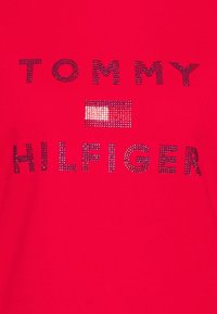 Punainen paita, jossa mustista strasseista muodostuva "TOMMY HILFIGER"-teksti ja pieni lippukuvio. Sileä, istuva kangaspinta.
