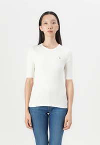 White ribbed short-sleeve top with a round neckline and small logo accent on the chest, paired with blue fitted jeans. Smooth texture.