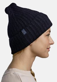 Woman in profile wearing a black ribbed knit beanie and light-colored shirt, smiling gently against a plain background.