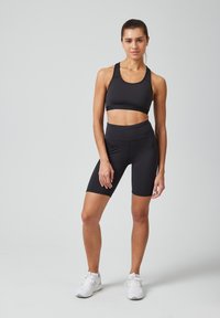 Zwarte sportbra en hooggerafelde fietsshorts van rekbaar materiaal; model staat in een neutrale omgeving en draagt witte sportschoenen.