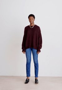 Suéter de punto burdeos, de corte relajado con mangas anchas, combinado con jeans ajustados azules y mocasines de tacón negros, de pie contra un fondo neutro.