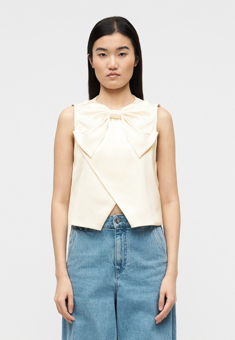 Γυναίκα με ίσια σκούρα μαλλιά φορά ει αμάνικο κρεμ τοπ με μεγάλο φιόγκο και ψηλόμεσο μπλε τζιν, στέκεται μπροστά σε απλό λευκό φόντο.