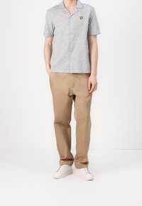 Graues Kurzarm-Hemd mit Knöpfen, feiner Textur und gesticktem gelben Logo, kombiniert mit beigen Hosen und weißen Turnschuhen.