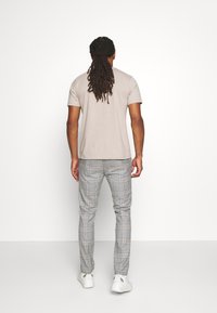 Muž s dlouhými dredy, oblečený v béžové košili s krátkým rukávem, šedých kostkovaných kalhotách a bílých teniskách, stojí čelem k bílé zdi.