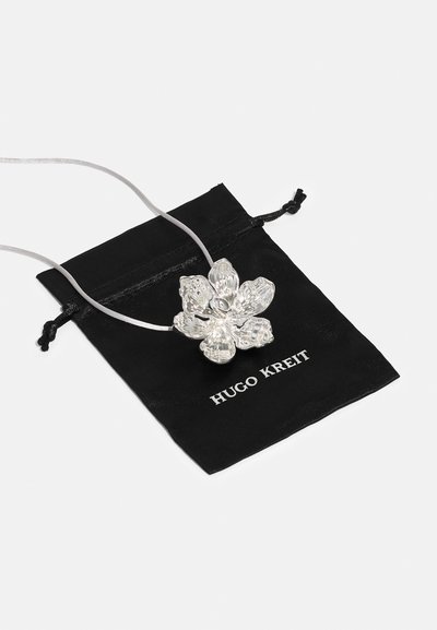 Hõbedane lille ripats peenel hallil nööril, resting mustal kotil, millel on valge tekst "HUGO KREIT". Tekstuuritud lillemustriga detail.